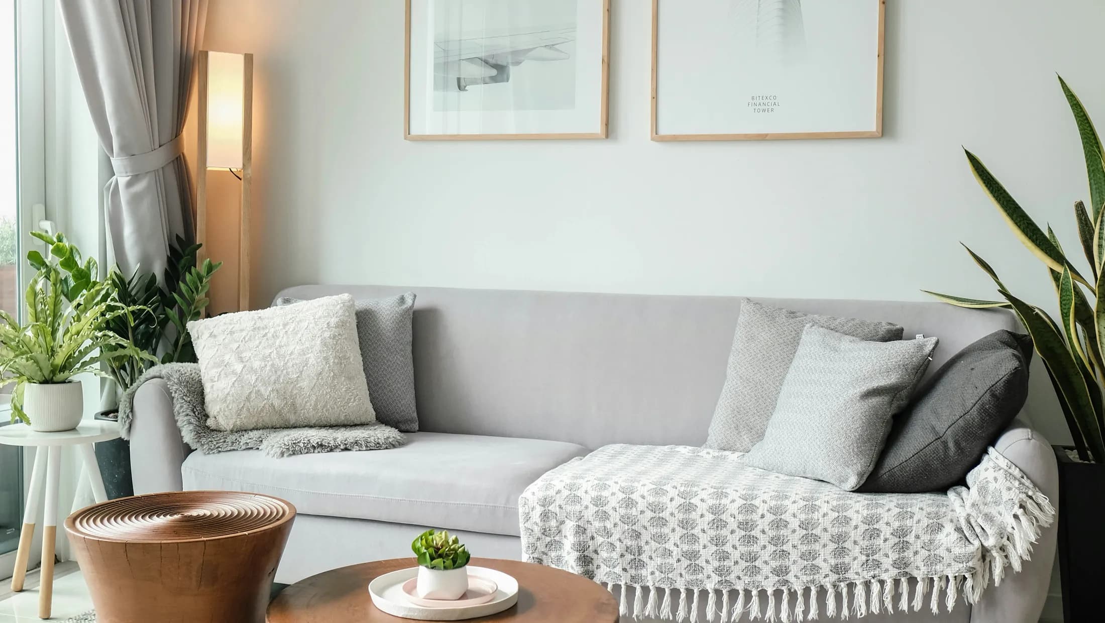 Pale grey sofa layered with textured cushions and a patterned throw, framed prints above and potted plants on either side