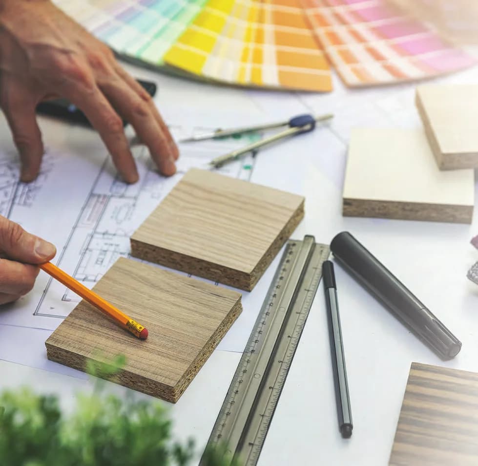 Hands with a pencil over a floor plan, surrounded by timber veneer samples, a colour fan, a compass and a ruler